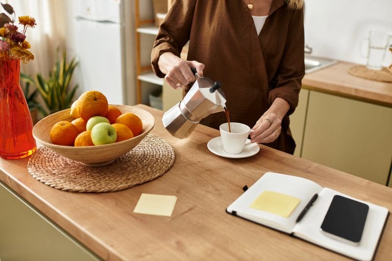 Photograph of a woman pouring coffee in her well-organized kitchen, with a planner, pen, smartphone, and post-it notes close at hand