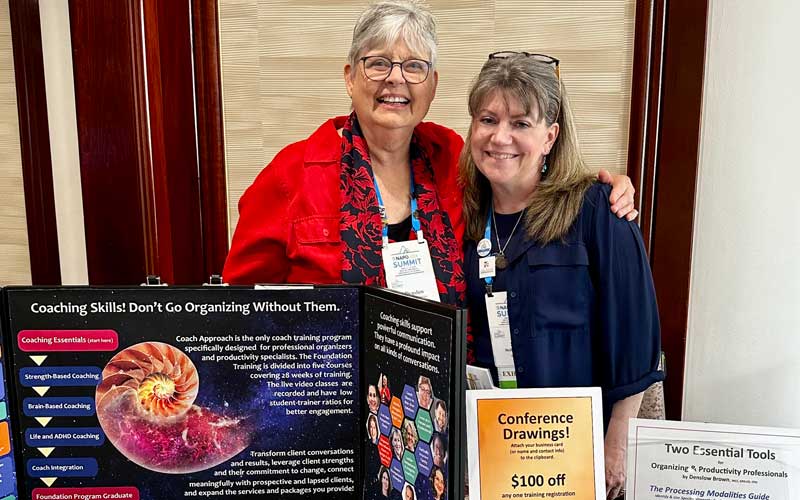 A photo of Denslow Brown and Sara Skillen at the Coach Approach Training table at the 2024 NAPO Summit. Display includes posters with the headings "Coaching Skills! Don't Go Organizing Without Them", "Conference Drawings!", "Two Essential Tools for Organizing & Productivity Professionals by Denslow Brown", and "Coaching skills support powerful communication. They have a profound impact on all kinds of conversations."